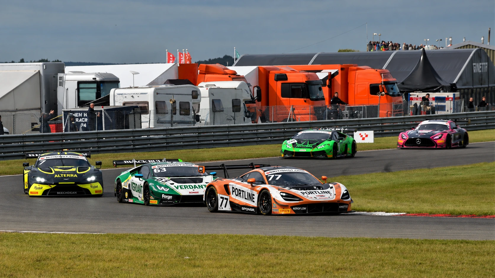 A British GT field streaming through a corner, a McLaren leading a Lamborghini, team transporters in the paddock behind (SPONSR livery concept).