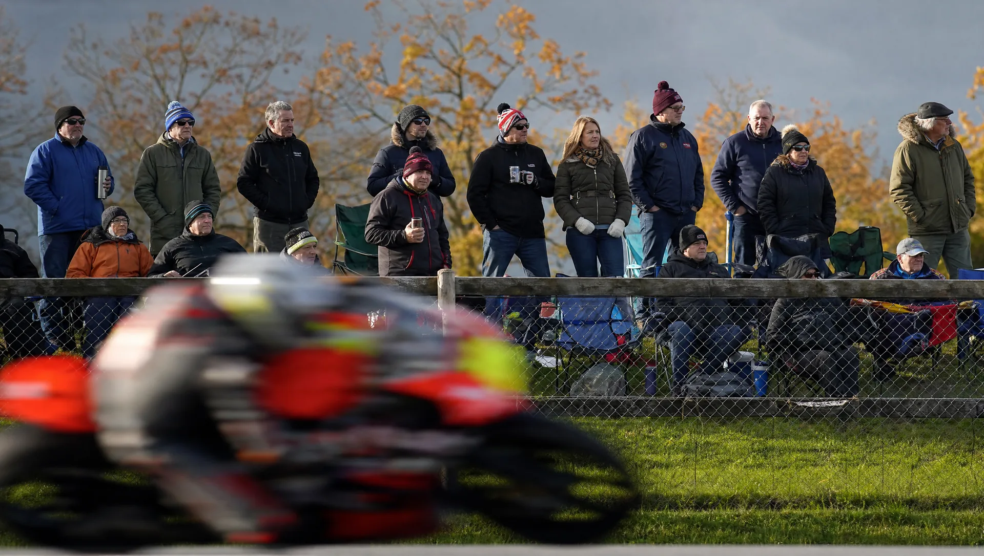 Families and friends along the spectator fence on a grass bank as a racing bike sweeps past below.