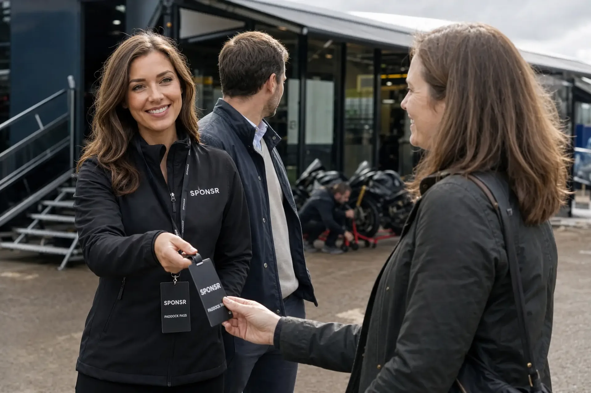 A SPONSR host handing a black paddock pass on a lanyard to a guest in the superbike paddock, two superbikes on stands under an awning behind them.